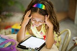 Girl wearing a rainbow headband sitting and looking and down at a tablet device.