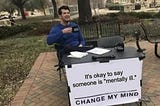 Man in a park sitting at a table with a sign in front saying “It’s okay to say someone is “mentally ill.” Change my mind.