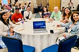Eight people sit at a roundtable with an open laptop.