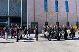 Two groups of percussionists face each other, one group carrying tall speakers on their backs, outdoors in front of a Theatre.