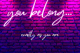 Image of a pink, purple, and blue lit brick wall with neon text reading “you belong… exactly as you are”