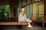 A woman with light brown hair is wrapped in a sweater and sitting cross-legged on a wooden bench.