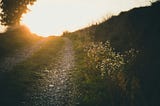 An unpaved path headed up a hill with the sun breaking ahead over the top