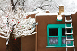 Color photo of a snowy winter scene showing a snow covered tree with christmas decorations in front of an adobe home.
