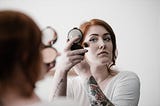 Woman looking into a mirror while applying makeup to her cheek.