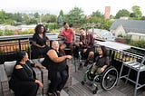 Overhead shot of six disabled people of color at a rooftop deck party. An Indigenous Two-Spirit person with a prosthetic leg smiles directly at the camera and gives a thumbs up while everyone else is engaged in conversation.