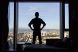 The silhouette of a man with his back turned standing at his full height with his hands on his hips, in front of a floor-to-ceiling window with a city view, with a chaise lounge sofa and an open laptop on a side table behind him.