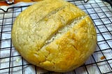 A round loaf of bread with three slits on the top is sitting on a baking rack cooking over a piece of parchment paper.