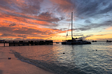 Large catamaran sailboat approaching pier on the beach at sunset.