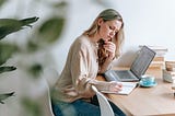 Woman writing at a desk with a laptop, notebook, and coffee.