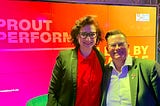 Two individuals stand smiling in front of a vibrant backdrop at the 2022 *Prout Performer* Networking Event, hosted by the Prout at Work Stiftung. On the left is Ferda Ataman, Germany’s Federal Anti-Discrimination Commissioner, advocating for equality and combating discrimination nationwide. On the right, Jay Siegmann, a *Prout Performer* Top 10 in 2022 and Top 5 in 2021, celebrates their contributions to LGBTQIA+ visibility and workplace inclusion.