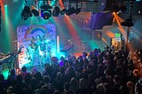 A photo of Madeline the Person performing in Seattle, Washington. Colorful rainbow lights wash over the stage as she sings and strums a guitar in front of a collaged backdrop of her and her fans art.