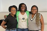 Three black women, with their arms around each other, stand side by side and are smiling to camera.