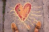 A red heart with yellow lines radiating from it draw in chalk on pavement with person’s shoes visible at the bottom of the frame.