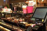 A two-level counter and display with sprinkled donuts, bright pink cups, and other gift items from Dunkin along with a cash register