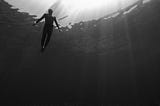 Person in water, seen from underneath. Their head is at the surface and their feet are well above the ocean floor. Black and white, with rays of light shining from above.