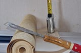 A coloured image showing a roll of wallpaper, a tape measure and a wallpaper scraper sitting on a metal bench against a bare wall background.
