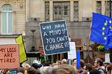 Photo of a protest. There people holding up signs which read (from left to right): “My legs are tired” and “Why have a mind if you can’t change it?”