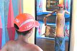 Young boy, around 8–9 years old wearing a red ball cap and jeans but no t-shirt standing in front of a funhouse mirror that stretches his torso way out and makes it look like he has three belly buttons