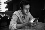 A man sitting at a table looks at a mobile phone that he’s holding in his hands.