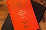 A vibrant orange book cover with the text “John Mark Comer. The Ruthless Elimination of Hurry. Forward by John Ortberg” sits on top of a black notebook on a wooden-look desk.