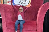 Librarian sitting on an oversized chair holding a sign saying, “Let Freedom Read!”