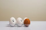 Three white eggs with faces drawn on them looking at a dark brown egg.