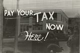 Black and white photo (1940's?) showing a window sign that says “Pay Your Tax Now Here!) with a street scene reflected in the glass behind