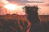 Warm orange and pink silhouette of a woman wearing a floral wreath, standing in a cornfield at sunset.