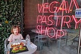 Child opens her mouth in awe about the delicious-looking vegan brunch at Gia’s Vegan Pastries in San José, Costa Rica. Light-up sign reads “VEGAN PASTRY SHOP.”