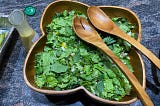 Mixed green salad in clover-leaf wooden bowl with wooden serving utensils and lemon dressing in a pouring container
