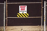 A cat sits behind a fence, peering through a gate with a sign that warns “keep out”