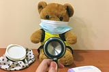 Photograph of brown teddy bear wearing a blue surgical mask while a doctor’s hands hold a stethoscope in front of it.