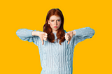 Red haired young woman in a light blue jumper with her arms raised in front of her, with her two thumbs pointing down. She looks a bit negative. Bright yellow background