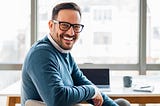 A writer smiling while sitting at a wooden desk. In the background, a laptop, coffee mug, and notebook are visible.