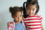 A Black child and Asian child facing the camera in a partial embrace against an off-white background. The Black child is a little girl with pigtails, wearing a blue shirt with a pink-and-white stripped cardigan. The taller Asian child is also wearing pigtails and is wearing a red-and-white stripped shirt.