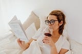 Woman in White Shirt Holding a Glass of Wine reading a book in bed.