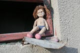 Kill, Let Kill, Or Be Killed. A small ragdoll sits abandoned, unclothed, and dirty in the corner of a glassless window with broken concrete around the pane.