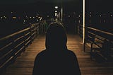A person wearing a hoodie in a shadow, standing on a boardwalk at night.