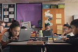 A group of elementary students in a classroom looking at laptops in their desks.