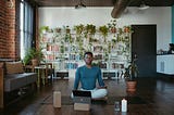 Man meditating in a room filled with books and plants and a burning candle.