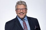 Profile picture of Wynn Hausser, a smiling middle-aged White man with glasses and a goatee.