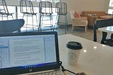 A small laptop opened to a writing program sitting on a coffee shop’s table. There is a to coffee to-go cup on the table.