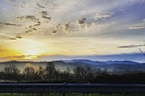 Over a metal railing, a pale yellow Appalachian sun rises over the Blue ridge Mountains, seen in the distance. Its cold, and the fog is thick. The stratus clouds are scarce, and feathered. It makes me sad.