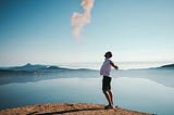 man wearing white shirt standing on top of hill overlooking ocean spreading his arms with face looking up to the sky