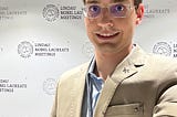 Selfie de Gustavo Rosa Gameiro, ele é um homem branco jovem, usa óculos, paletó beje e camisa azul e ao fundo está uma parede com o marca e texto do “Lindau Nobel Laureate Meetings”