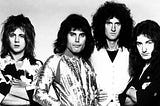 Roger Taylor, Freddie Mercury, Brian May, and John Deacon of the band Queen photographed in 1975, wearing glam outfits and teased hairdos, looking directly at the camera