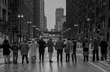 A black and white photo of about 10 people linked arm-in-arm across a city street. The street is slick with a recent rain, and some people in the photo are wearing rain gear and ponchos. The city rises around them with street lights and tall buildings in the background.