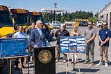 Photo of Gov. Inslee standing outdoors at a podium with a small group of people behind him. Behind the group are new electric school buses, and next to the podium are two posters — one showing increasing oil company profits and another showing a collage of headlines about oil company profits and climate-related weather events and disasters.