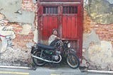 Old brick wall and reddish brown wooden door, in front of it is a motorcycle leaning against the door, a painting of a boy appears to be sitting on the motorcycle and looking over his shoulder at a line drawing of a dinosaur